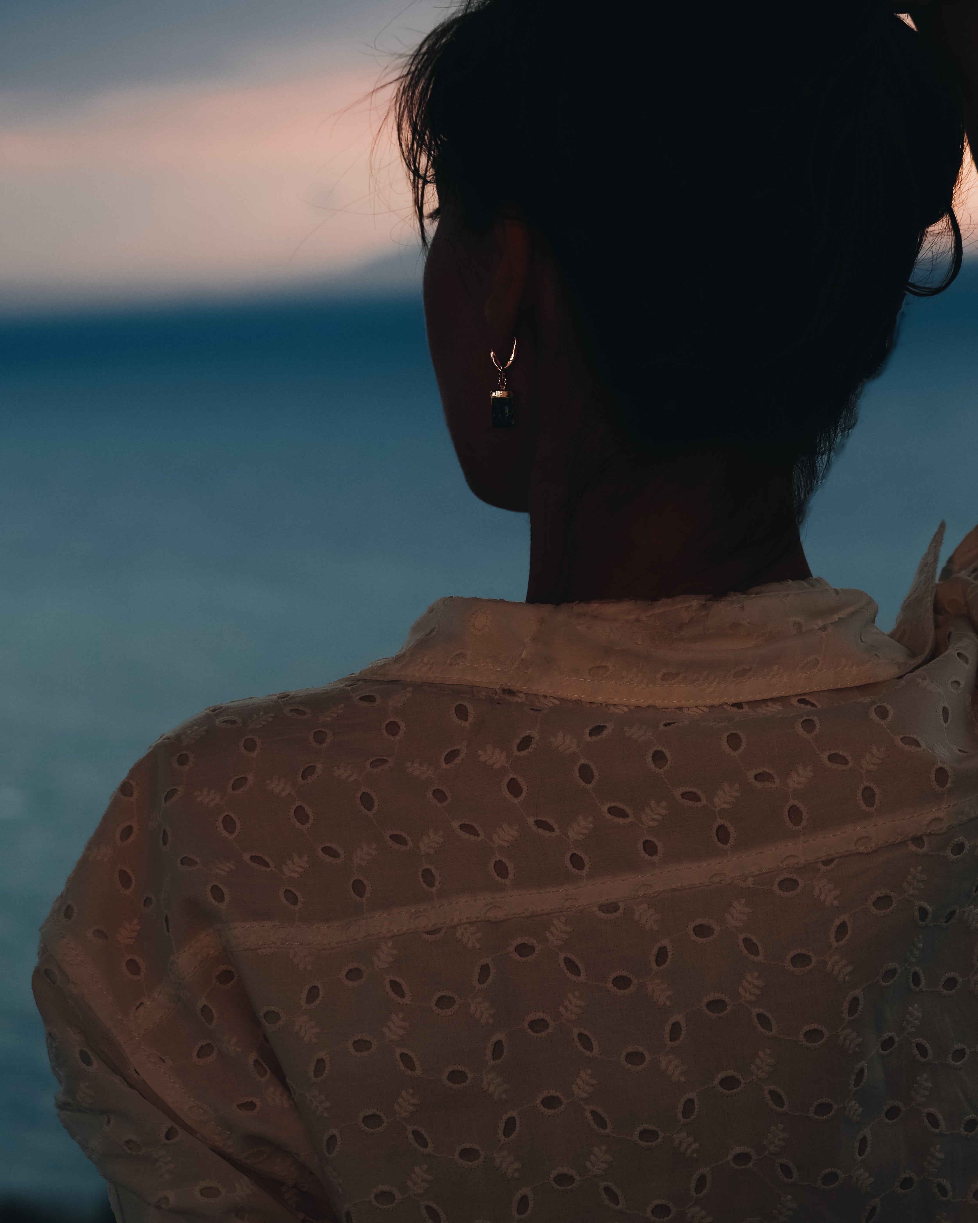 Une femme portant une boucle d’oreille élégante regarde l’océan depuis les hauteurs au coucher du soleil — une vision de beauté intérieure, de force tranquille et d’harmonie avec le monde.