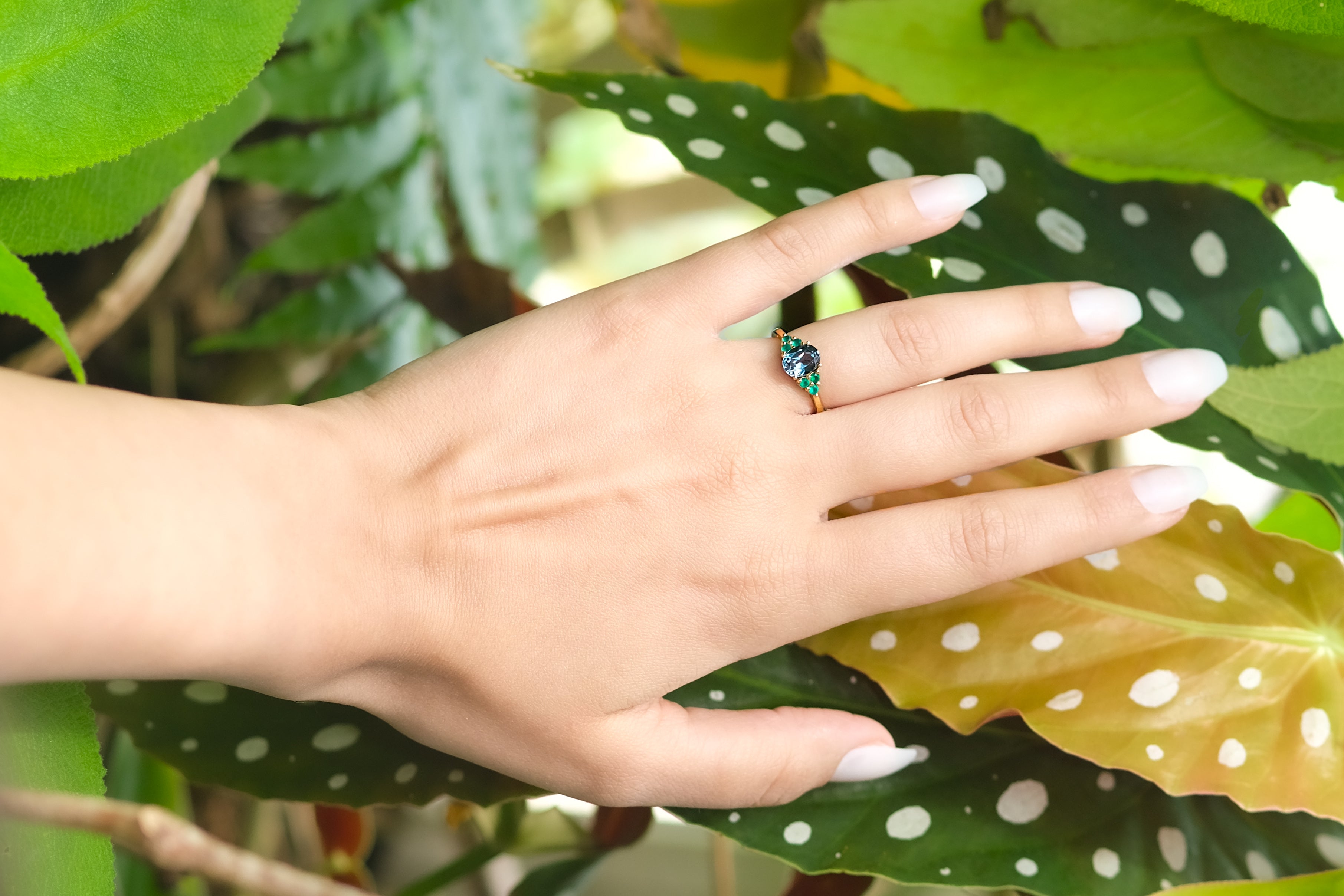 Une main portant la bague Blue Velvet touche délicatement des feuilles vertes et jaunes mouchetées de blanc — une image d’élégance éthique et de lien avec la nature.
