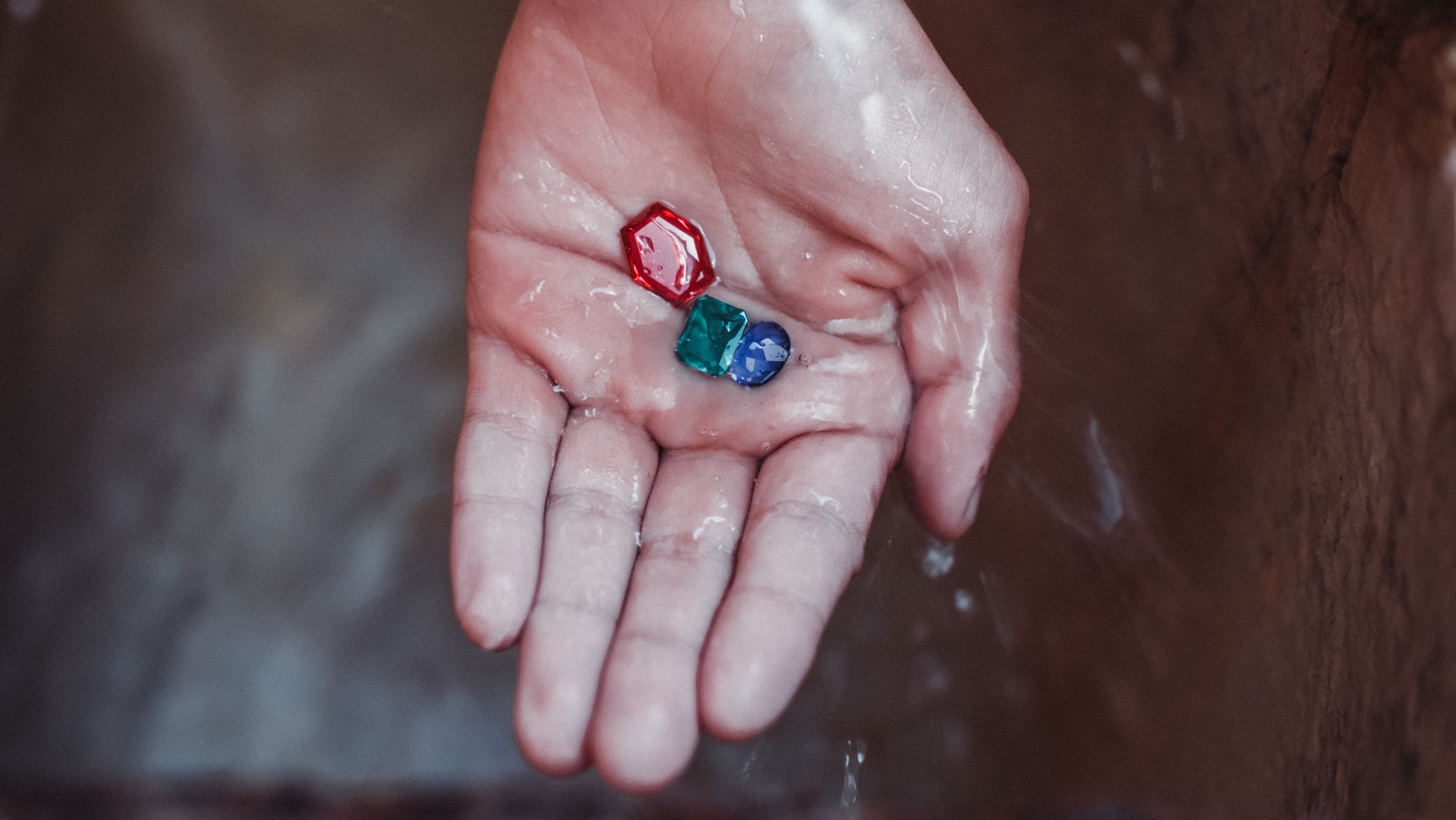 Une main ouverte recevant de l’eau claire tandis que des rubis, des émeraudes et des saphirs reposent dans sa paume — un symbole de pureté, de beauté consciente et de renouveau.